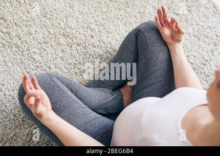 Vue au-dessus de la jeune femme enceinte dans des leggings assis avec des jambes croisées sur la moquette et tenant les mains dans la mudra tout en méditant à la maison Banque D'Images