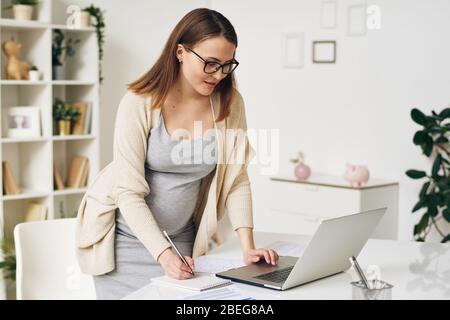 Femme enceinte moderne dans des lunettes et un gilet debout à la table et à l'aide d'un ordinateur portable tout en écrivant des tâches dans le bloc-notes Banque D'Images