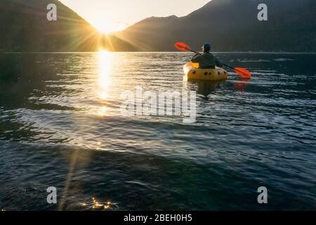 Un jeune homme pagayant sur le lac au coucher du soleil vers les montagnes Banque D'Images