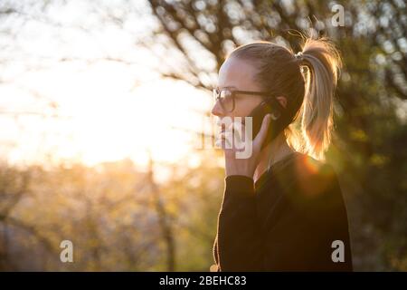 Vue arrière rétroéclairé de la jeune femme parlant sur le téléphone portable à l'extérieur dans le parc au coucher du soleil. Fille tenant le téléphone mobile, à l'aide de l'appareil numérique, regarder le réglage Banque D'Images