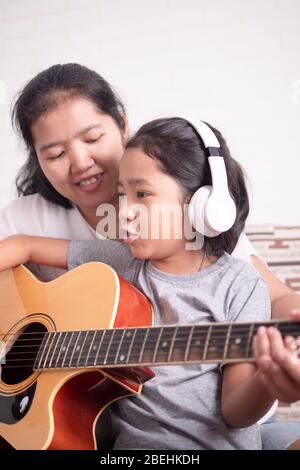 Maman enseigne à sa fille à jouer de la guitare. Petite fille asiatique apprenant à jouer de la musique et à chanter une chanson. Les enfants portent un casque blanc. Banque D'Images