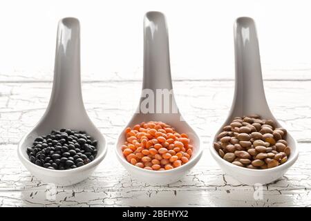 Différents lentilles assorties se mélangent avec des lentilles béluga rouges, brunes et noires dans des cuillères blanches sur fond de table en bois blanc - foyer sélectif Banque D'Images