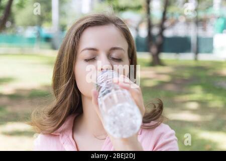 Jeune femme caucasienne fille boire de l'eau d'une bouteille en plastique dans le parc vert, gros plan Banque D'Images