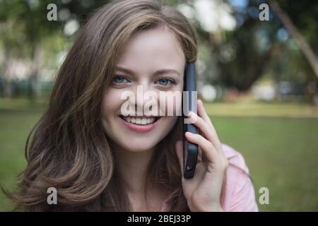 Jeune fille blonde souriante caucasienne parlant au téléphone dans le parc Banque D'Images