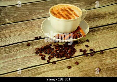 Une tasse blanche avec cappuccino frais saupoudrée sur une soucoupe décorée de bâtonnets de cannelle et de chandelles sur une table en bois dans une pile de grains de café. C Banque D'Images