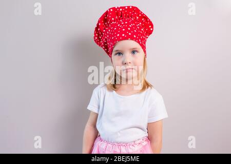 Petite fille blonde avec un visage sérieux dans le chapeau de cuisinier rouge restant sur un fond gris neutre. Banque D'Images