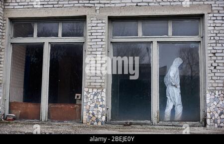 Pleine longueur de l'environnement masculin debout à l'intérieur de la vieille maison en brique avec de grandes fenêtres. Un scientifique portant un costume de protection blanc et un masque à gaz tout en explorant les problèmes environnementaux à la centrale électrique. Banque D'Images