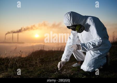 Scientifique en costume de protection et masque à gaz prenant l'échantillon de sol à la centrale électrique. Chercheur avec cheminées thermiques fumeurs et beau ciel sur fond. Concept de recherche et d'écologie. Banque D'Images