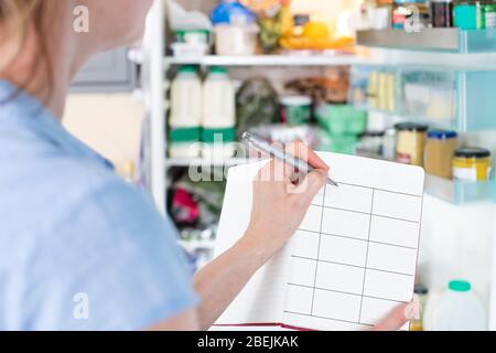 Femme debout devant le réfrigérateur dans la cuisine avec carnet écriture repas hebdomadaire Banque D'Images