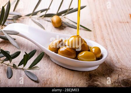 L'huile d'olive extra vierge s'écoule dans le bol en céramique rempli d'olives vertes sur la table en bois rustique Banque D'Images