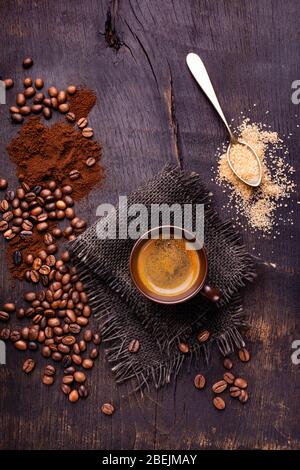 Sur la table en bois brut et sombre, une tasse de café avec une cuillerée de sucre brun et de grains de café torréfiés. Banque D'Images