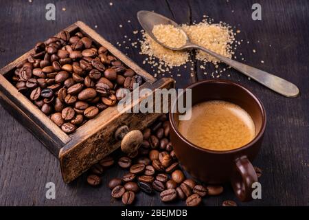 Sur la table en bois brut et sombre, une tasse de café avec une cuillerée de sucre brun et de grains de café torréfiés. Banque D'Images