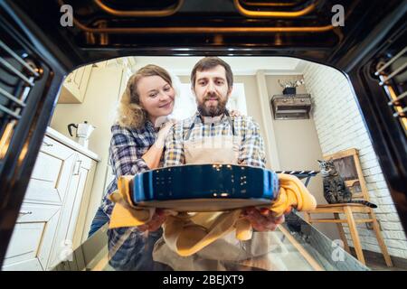 Couple souriant d'Europe de l'est cuisine à la maison. Homme sortant une tarte du four avec cuisine intérieure et un chat sur fond, vue de l'intérieur de th Banque D'Images