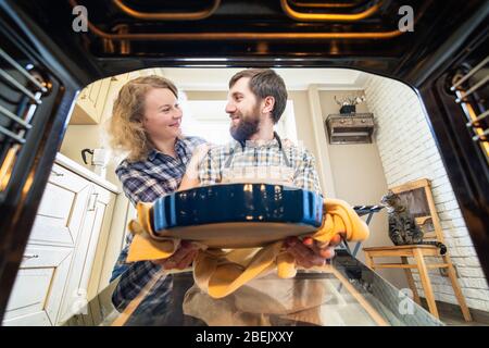 Couple d'Europe de l'est cuisine à la maison, souriant et regardant l'un l'autre. Homme sortant une tarte du four avec cuisine intérieure et un chat à l'arrière Banque D'Images