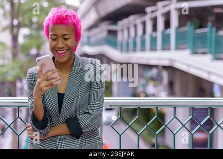 Heureuse jeune femme d'affaires rebelle d'Asie avec des cheveux roses utilisant le téléphone dans la ville Banque D'Images
