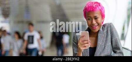Heureuse jeune femme d'affaires rebelle d'Asie avec des cheveux roses utilisant le téléphone dans la ville Banque D'Images
