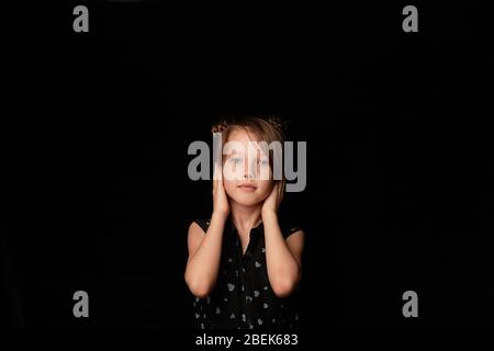Une fille de 6 ans, sur un fond sombre.couvre ses oreilles avec ses mains. Je n'entends pas le mal. Banque D'Images