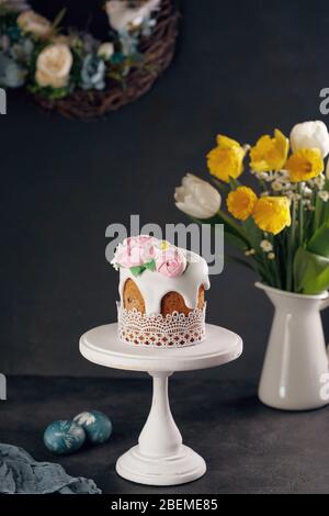 Gâteau de Pâques fait maison sur un stand en bois blanc avec bouquet de jonquilles et tulipes dans un vase sur fond sombre. Concept de décoration de vacances. Banque D'Images