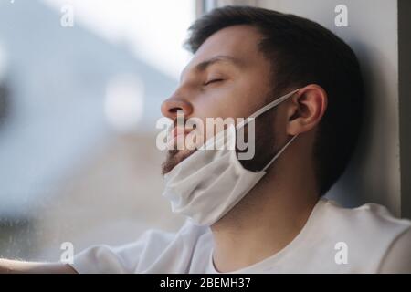 Jeune homme assis sur le seuil de la fenêtre et tiré le masque de protection sous le menton et regarder hors de la fenêtre. Seul à la maison en quarantaine. Thème du coronavirus Banque D'Images