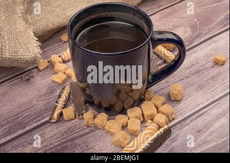 Mug en céramique bleu foncé avec thé, bâtonnets de biscuit avec chocolat et glaçage blanc et morceaux de sucre de canne brun sur fond en bois. Gros plan Banque D'Images