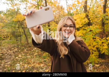 Belle jeune femme gaie portant un manteau marchant dans le parc d'automne, prenant un selfie Banque D'Images