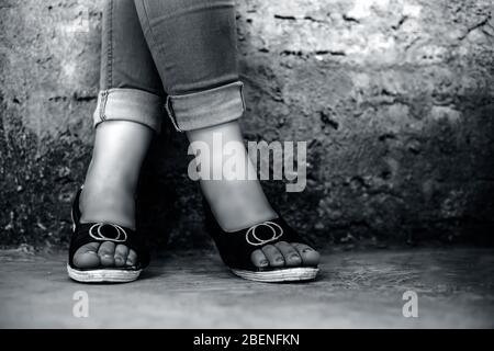 Photo du pied d'une jeune fille élégante portant un Jean denim bleu et des sandales à bout ouvert posant des jambes croisées contre le fond de mur de briques en noir et blanc Banque D'Images