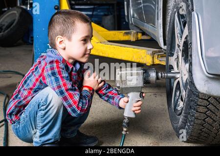 Le garçon rêve de devenir adulte et de réparer des voitures. Un jeune garçon, un jeune travailleur automobile, fait un changement de pneu avec une clé pneumatique dans le garage de Banque D'Images