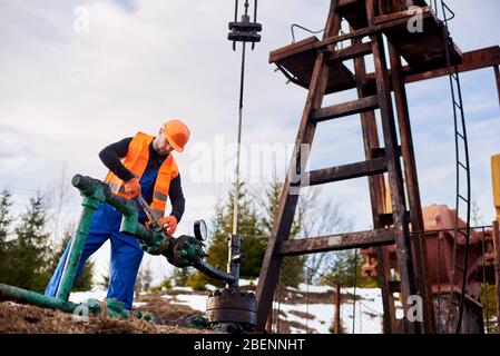 Ouvrier du pétrole dans les combinaisons bleues, gilet orange et casque travaillant avec un boulon sur un tuyau à gaz près d'un cric de pompe à huile, concept de l'industrie pétrolière Banque D'Images