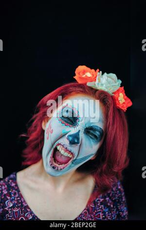 Jeune femme dans le jour du crâne masque mort visage art et cheveux rouges, criant sur fond sombre Banque D'Images