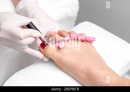 Les mains peignent du vernis à ongles rouge sur les orteils de la femme portant des gants blancs. Concept de soin des ongles et de pédicure. Banque D'Images