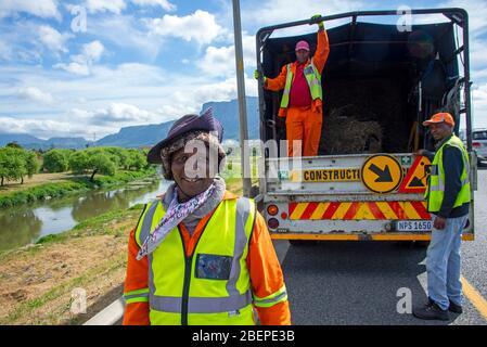 Trois travailleurs noirs du secteur public se posent sur le côté d'une autoroute où ils coupent le bord de l'herbe. Ils sont heureux et bien habillés dans des couleurs vives. La route se trouve à la périphérie de Cape Town, en Afrique du Sud. L'homme le plus proche présente des coiffures distinctives. Banque D'Images