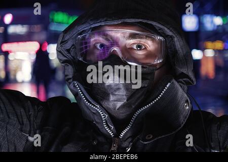 Homme sérieux dans les lunettes de sécurité, le masque médical et la capuche contre la rue éclairée. Portrait de type selfie Banque D'Images