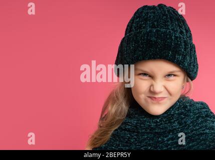 Petit enfant blond habillé d'un foulard et d'un chapeau chauds. Elle souriait, elle a l'air méchante, se posant sur fond de studio rose Banque D'Images