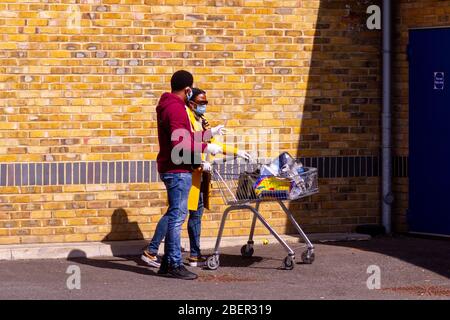 Homerton, Londres, Royaume-Uni, 7 avril 2020: Deux personnes en attente dans la file d'attente avec des sacs de trolley et de shopping ainsi que des masques et des gants de visage Banque D'Images