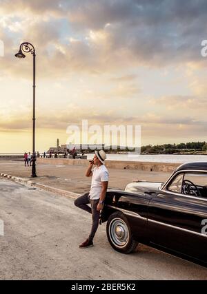 Cigare modèle cubain fumage par sa voiture Chevrolet d'époque à El Malecon, Château El Morro en arrière-plan, la Havane, la province de la Habana, Cuba (MR) Banque D'Images