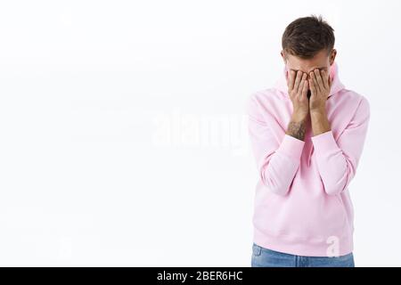 Un gars humilié couvre le visage avec les mains. Portrait d'homme blond mal facile dans le sweat à capuche rose, soupirer se sentir fatigué et affligé, ayant l'épuisement émotionnel Banque D'Images