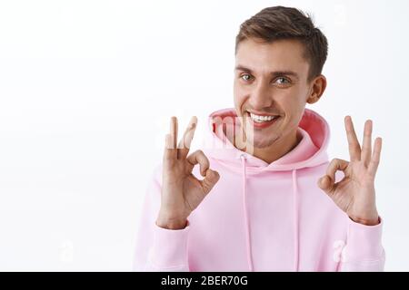 Satisfait, bon look blond jeune homme dans le hoodie rose, tout comme votre grande idée, montrer bon geste, garantir la meilleure qualité, recommander le produit Banque D'Images