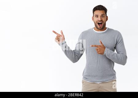 Un homme hispanique à la barbe, inspiré et enthousiaste, est un modèle élégant et gris qui survolez le t-shirt, pointant vers la gauche, souriant et joyeux Banque D'Images