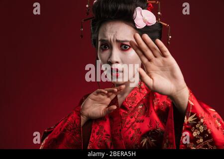 Image d'une femme géisha asiatique effrayée dans un kimono japonais traditionnel faisant un geste d'arrêt avec main isolée sur fond rouge Banque D'Images