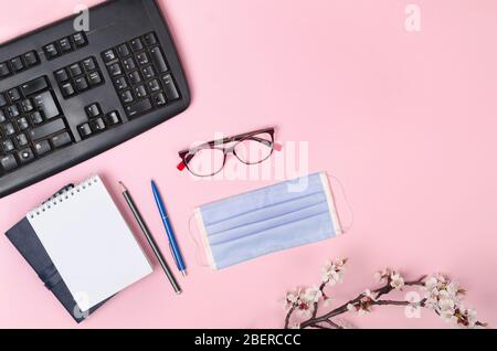 Branches fleuries avec un masque médical, un clavier, un bloc-notes et des lunettes sur un fond rose. Coronavirus COVID-19 Banque D'Images
