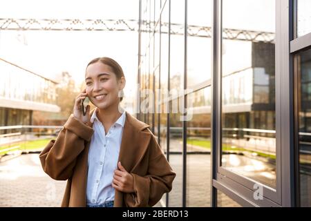 Image de la jeune femme asiatique brunette portant un manteau à l'aide d'un téléphone mobile tout en marchant dans la rue de la ville Banque D'Images