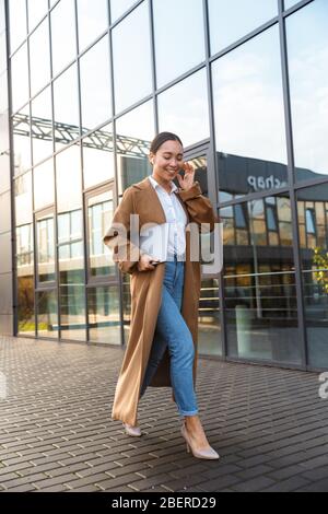 Image de la jeune femme asiatique brunette portant des écouteurs tenant un ordinateur portable tout en marchant dans la rue de la ville Banque D'Images