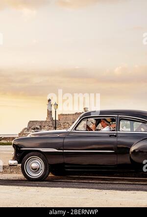 Cigare modèle cubain fumage dans sa voiture Chevrolet d'époque à El Malecon, Château El Morro en arrière-plan, la Havane, la province de la Habana, Cuba (MR) Banque D'Images