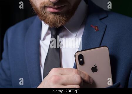 Zaporozhye. Ukraine. Février 2020. Gros plan portrait d'un jeune homme barbu dans un costume d'affaires tenant un iPhone XS dans sa main. Nouveau smartphone de Banque D'Images