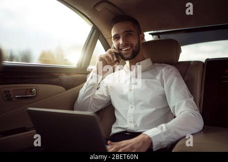L'homme utilise un ordinateur portable et parle sur un smartphone assis sur le siège arrière de la voiture, homme d'affaires en taxi Banque D'Images