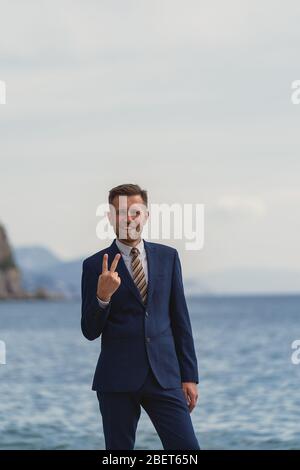 Homme d'affaires debout sur la plage en costume. Regarde l'appareil photo et souriant montrant le geste numéro deux Banque D'Images