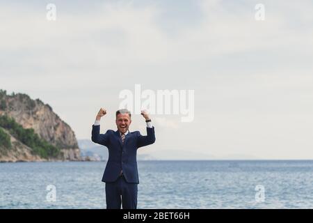 Homme d'affaires excité dans un costume criant et levant les mains au ciel par la mer Banque D'Images