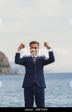 Homme d'affaires excité dans un costume criant et levant les mains au ciel par la mer Banque D'Images