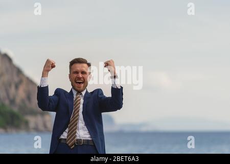 Homme d'affaires excité dans un costume criant et levant les mains au ciel par la mer Banque D'Images