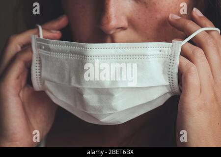 Photo détaillée d'une femme blanche non reconnaissable mettant sur un masque médical blanc pour empêcher la grippe. Fond flou, foyer sur le masque. Coronavirus, COVID-19 quarantaine. Concept médecin/infirmière. Banque D'Images